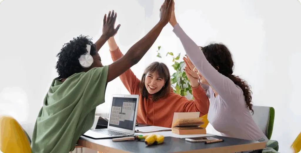 Diverse tech team high-fiving at a desk with a laptop, celebrating professional success.
