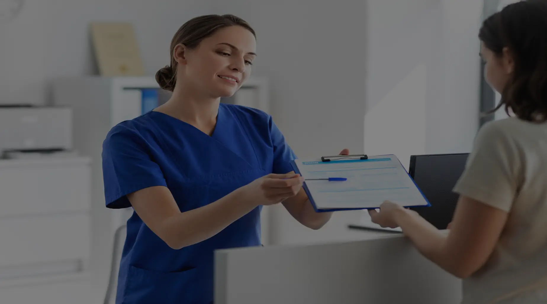 A female healthcare professional in blue scrubs handing a clipboard and pen to a patient at a medical reception desk.