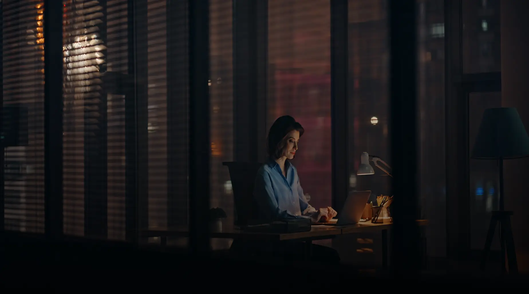 A woman working alone on a laptop at night in a dark office, illuminated by a desk lamp and city lights through the window.